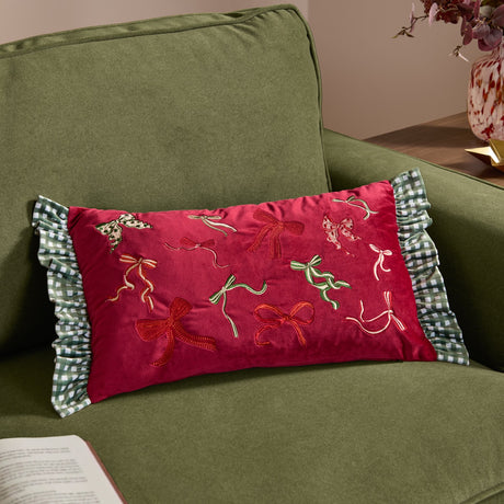 Rectangular red velvet Ditsy Bows cushion with embroidered bow designs and green-white gingham ruffle edges, styled on an olive green sofa.