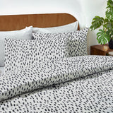 Dottie Printed Spotted Duvet Cover Set in black-on-white polycotton, shown on a bed with matching pillowcases against a wooden headboard in a bedroom setting.