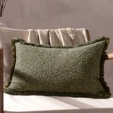 Doze Reversible Cotton Cushion in Moss, a rectangular fringed cushion with a flecked green boucle texture, shown on a chair with a folded throw against a neutral background.