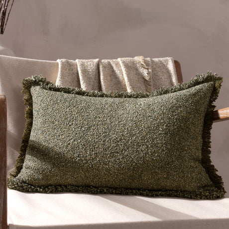 Doze Reversible Cotton Cushion in Moss, a rectangular fringed cushion with a flecked green boucle texture, shown on a chair with a folded throw against a neutral background.