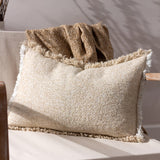 Doze Reversible Cotton Cushion in natural, a rectangular bolster with a textured cream and sandy beige weave and raw fringed trim, shown on a chair beside a woven throw.