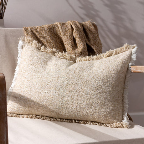 Doze Reversible Cotton Cushion in natural, a rectangular bolster with a textured cream and sandy beige weave and raw fringed trim, shown on a chair beside a woven throw.
