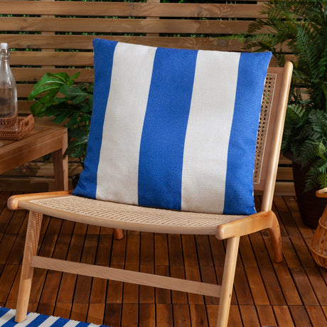 Deck Stripe reversible square cushion in bold blue and off-white polyester stripes, shown on a wooden outdoor chair against a slatted wood backdrop with greenery.