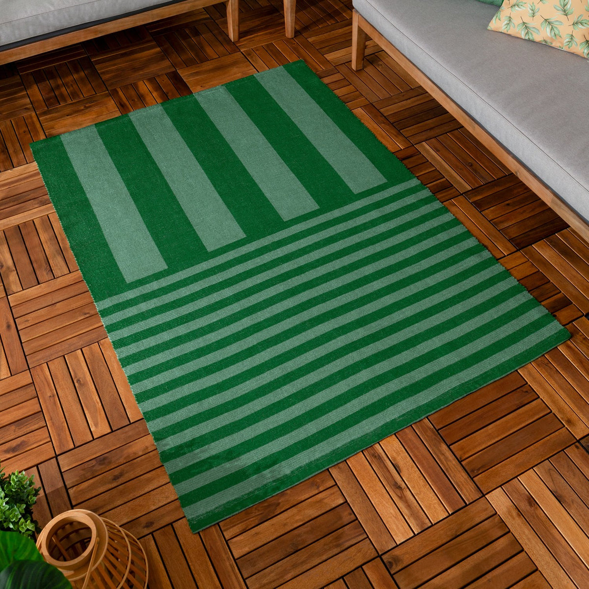 Deck Stripe Printed Polyester Rug in green, flatweave with broad vertical bars shifting to narrow horizontal stripes, shown on a wood-tiled floor beside a grey sofa.