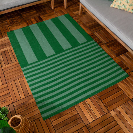 Deck Stripe Printed Polyester Rug in green, flatweave with broad vertical bars shifting to narrow horizontal stripes, shown on a wood-tiled floor beside a grey sofa.