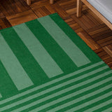Deck Stripe Printed Polyester Rug in green, flatweave with bold vertical bars shifting to narrow horizontal stripes at one end, shown on a wooden deck near patio furniture.