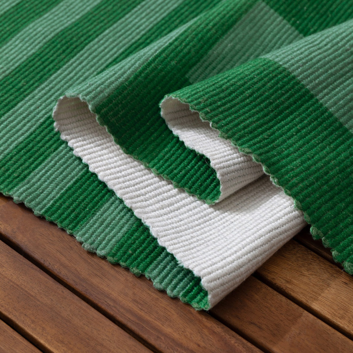 Deck Stripe Printed Polyester Rug in green, corner folded back to show white reverse side and ribbed flatweave texture, displayed on a wooden slatted surface.