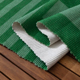 Deck Stripe Printed Polyester Rug in green, corner folded back to show white reverse side and ribbed flatweave texture, displayed on a wooden slatted surface.