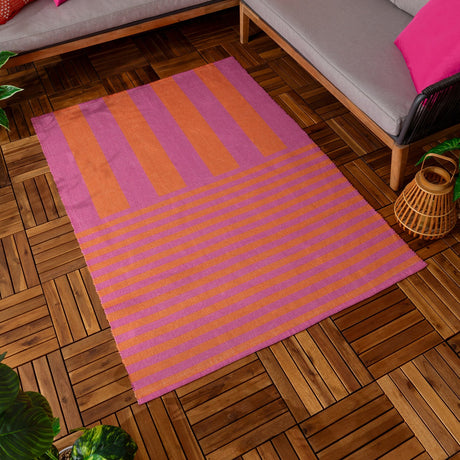 Deck Stripe Printed Polyester Rug in orange and pink, with vertical bands on the upper section and horizontal stripes below, laid on a wooden deck floor beside outdoor sofa seating.