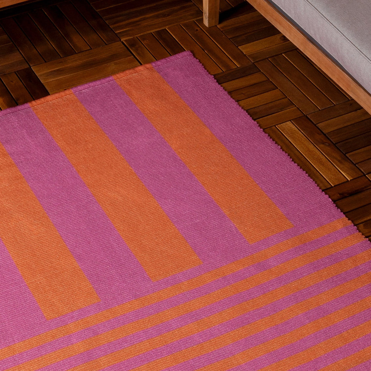 Deck Stripe Printed Polyester Rug in orange and pink, bold two-tone stripes shifting from wide vertical bands to narrow horizontal lines, shown on a wooden herringbone floor beside a sofa.