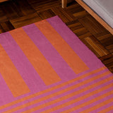 Deck Stripe Printed Polyester Rug in orange and pink, bold two-tone stripes shifting from wide vertical bands to narrow horizontal lines, shown on a wooden herringbone floor beside a sofa.