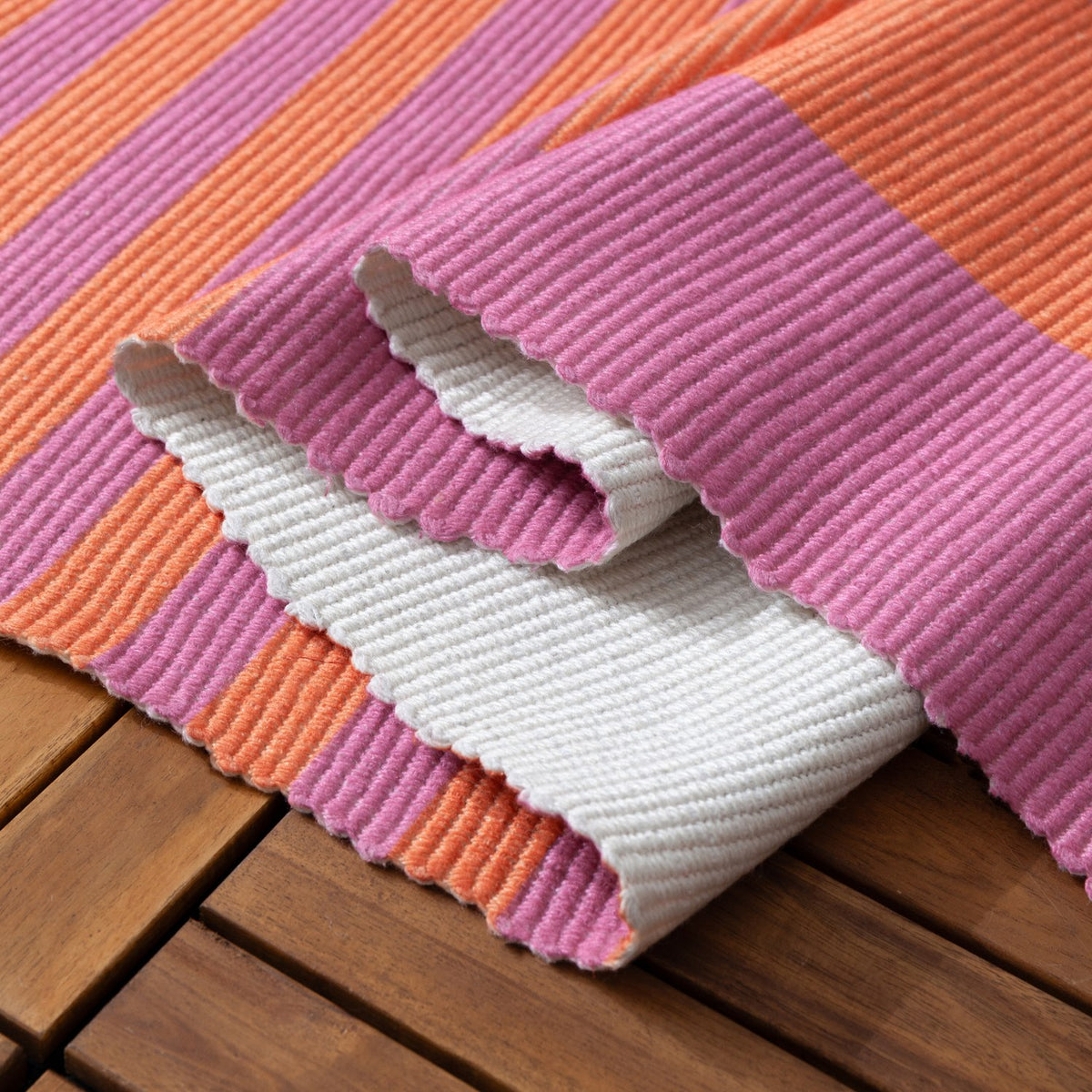 Deck Stripe Printed Polyester Rug in orange and pink stripes, corner folded back to show a plain white underside, laid on an outdoor wooden deck.
