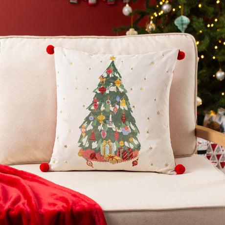 Deck The Halls square velvet cushion in ivory, featuring a printed Christmas tree with gold-embroidered ornaments and red pom-pom corners, shown on a living room armchair.