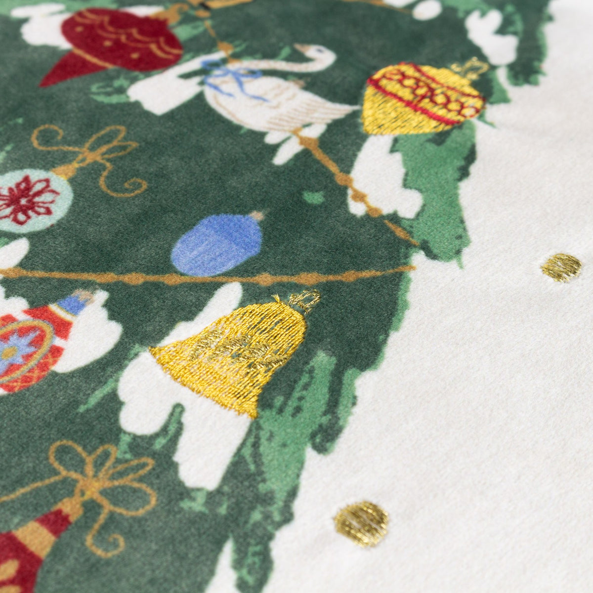 Close-up of the Deck The Halls velvet cushion, showing an ivory-white ground with green tree detail, red and blue baubles, gold embroidered bell, and scattered gold dot accents.