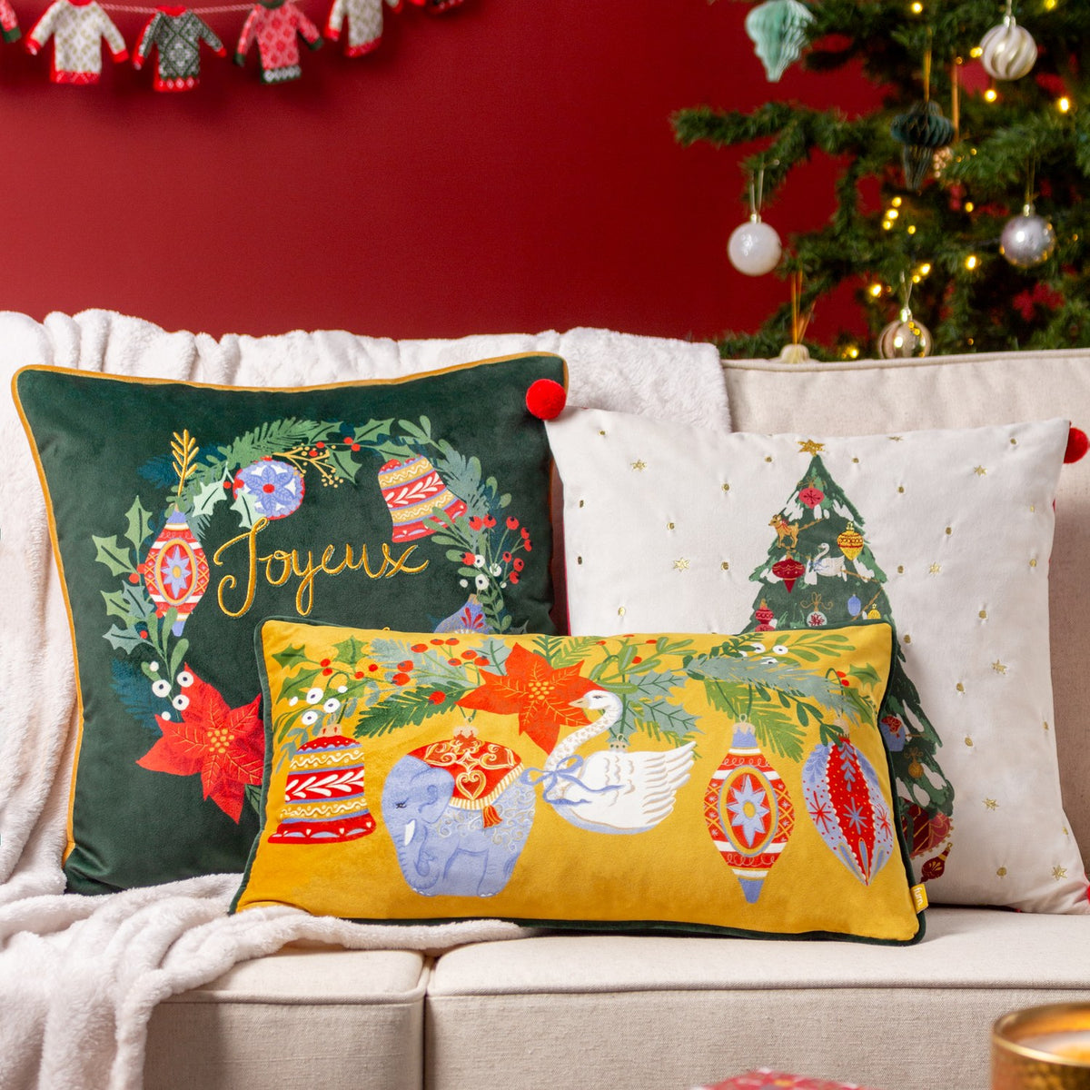 Velvet cushions on a sofa featuring a green wreath design with Joyeux embroidery, an ivory Christmas tree cushion, and a mustard bolster with festive ornaments and animals.