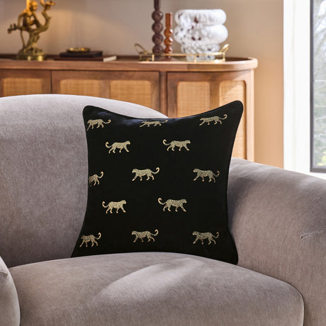 Duma Velvet Cotton Cushion in Noir, a black cotton velvet square cushion with embroidered gold cheetah motifs, shown on a grey armchair in a living room setting.
