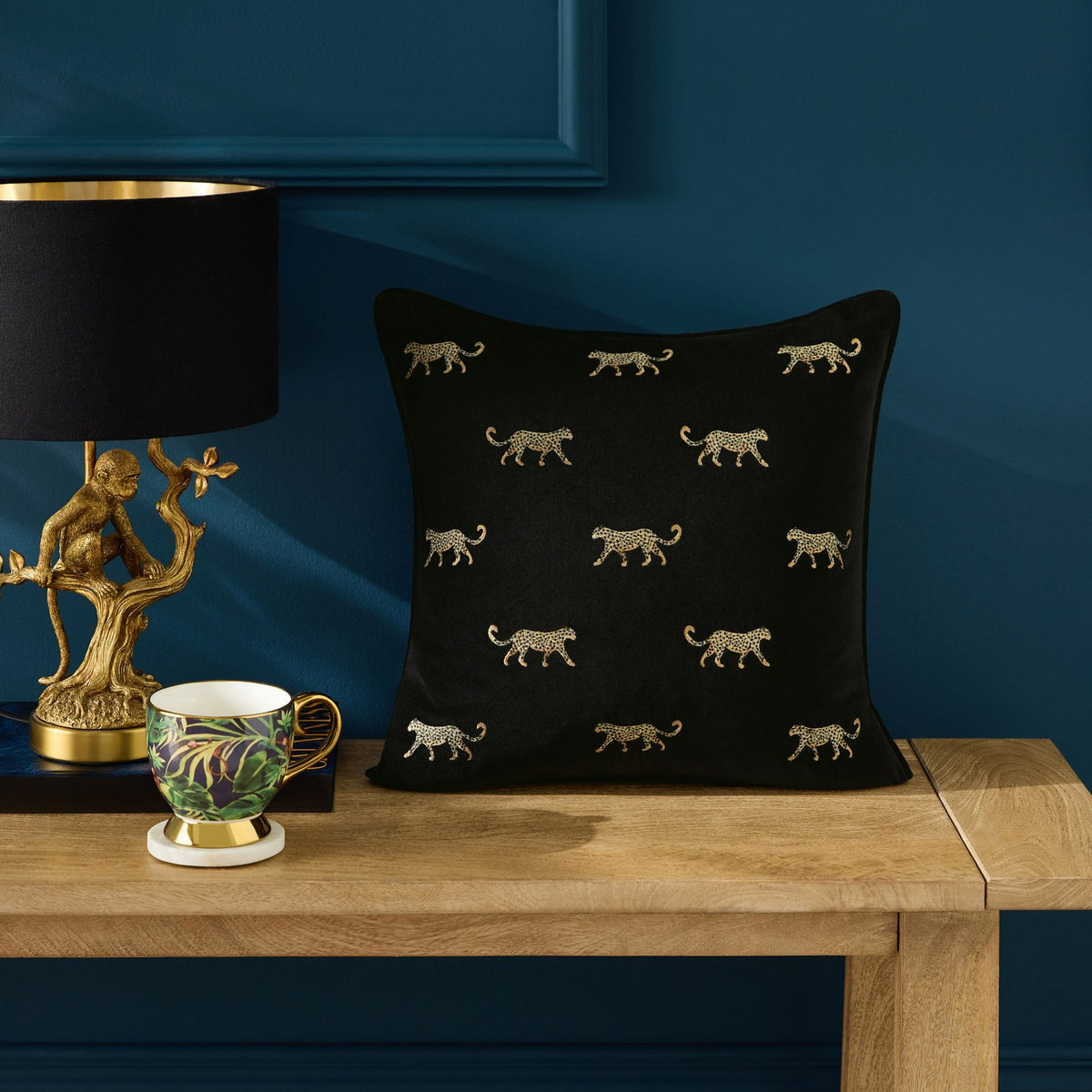 Duma Velvet Cotton Cushion in noir, black square cushion with gold embroidered cheetah motifs, shown on a wooden console table beside a patterned teacup and gold monkey lamp.
