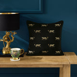 Duma Velvet Cotton Cushion in noir, black square cushion with gold embroidered cheetah motifs, shown on a wooden console table beside a patterned teacup and gold monkey lamp.