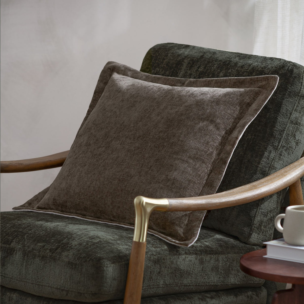 Dune Velvet Chenille Cushion in Mole, a square mid-brown chenille cushion with piped edging, shown on a dark green upholstered armchair with wooden armrests.
