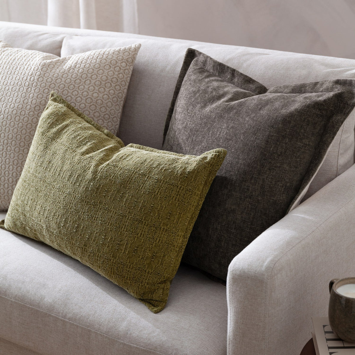Dune Velvet Chenille Cushion in Mole, a square mid-brown textured cushion with subtle sheen, styled on a light grey sofa with green and patterned cushions.