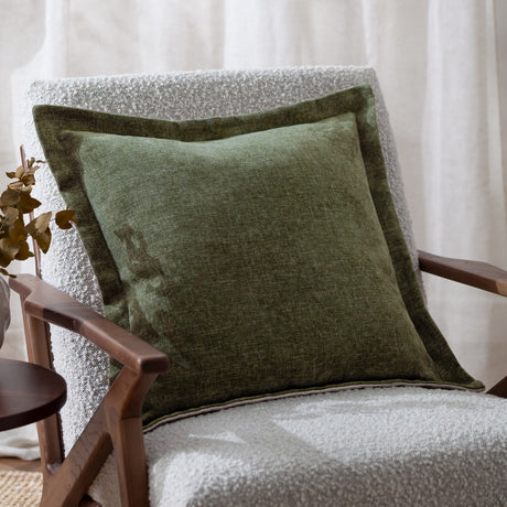 Dune Velvet Chenille Cushion in moss green with a wide oxford edge, shown on a bouclé armchair with a wooden frame.