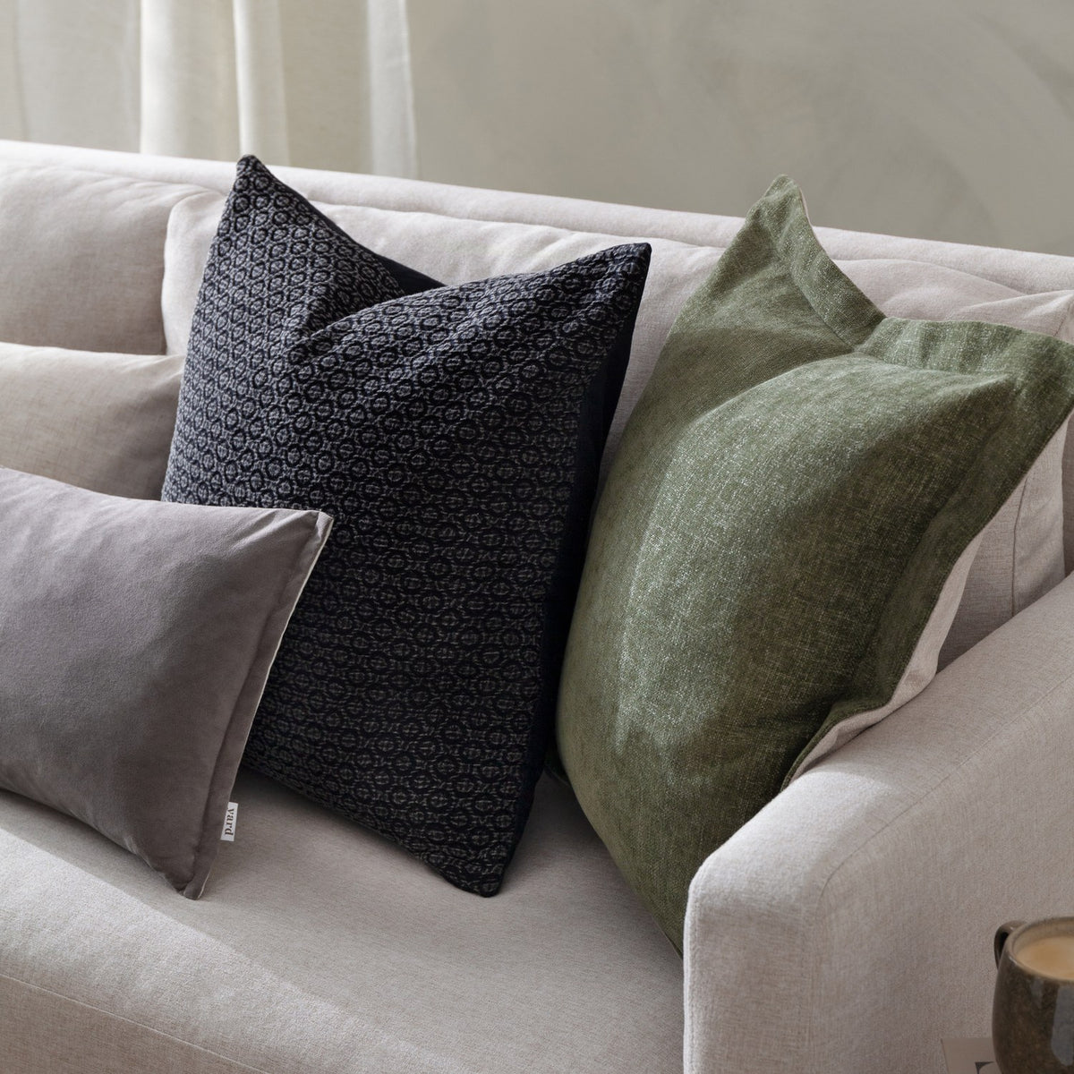 Dune Velvet Chenille Cushion in moss green with an oxford edge, styled on a beige sofa alongside black patterned and grey velvet cushions.