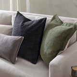 Dune Velvet Chenille Cushion in moss green with an oxford edge, styled on a beige sofa alongside black patterned and grey velvet cushions.