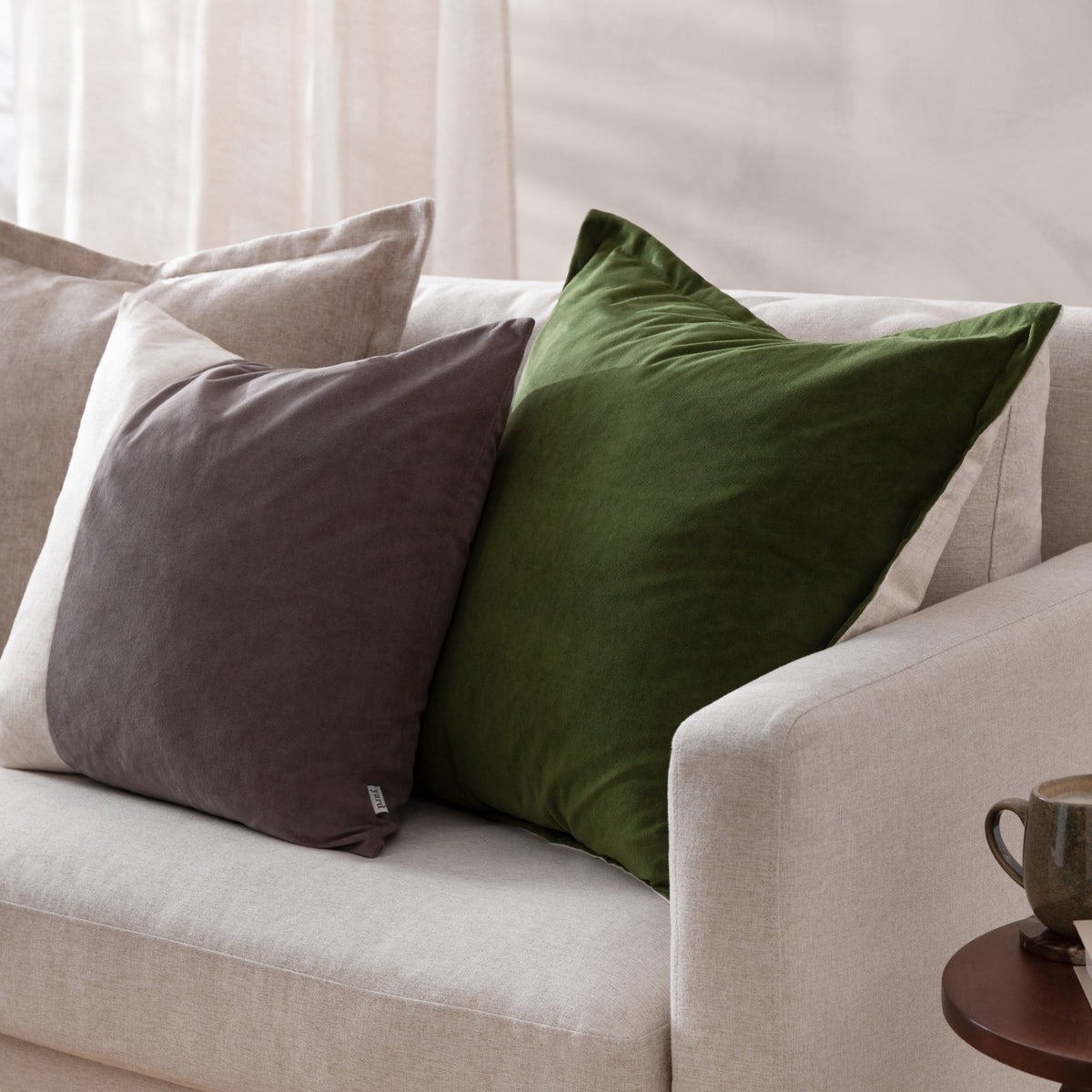 Dune Velvet Chenille Cushion in Natural, square with flat oxford border, shown on a beige sofa beside a green velvet cushion in a living room setting.