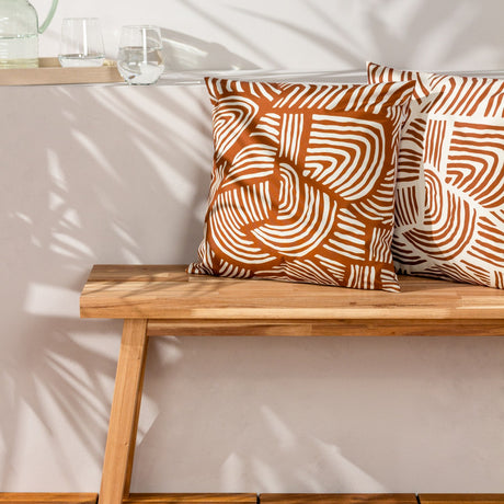 Dunes Reversible Polyester Cushion in Brick with cream swirling line pattern, shown on a wooden bench in a sunlit indoor setting.