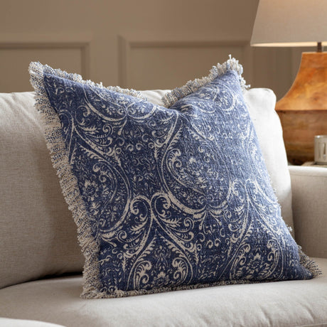 Durelle Damask Cotton Cushion in Delph Blue, square with raw fringed edge and scrolling floral pattern on cream cotton, shown on a beige sofa in a living room.