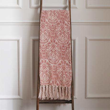 Durelle Damask Cotton Throw in coral pink and cream paisley pattern with knotted tassel fringe, draped over a wooden ladder against a panelled wall.