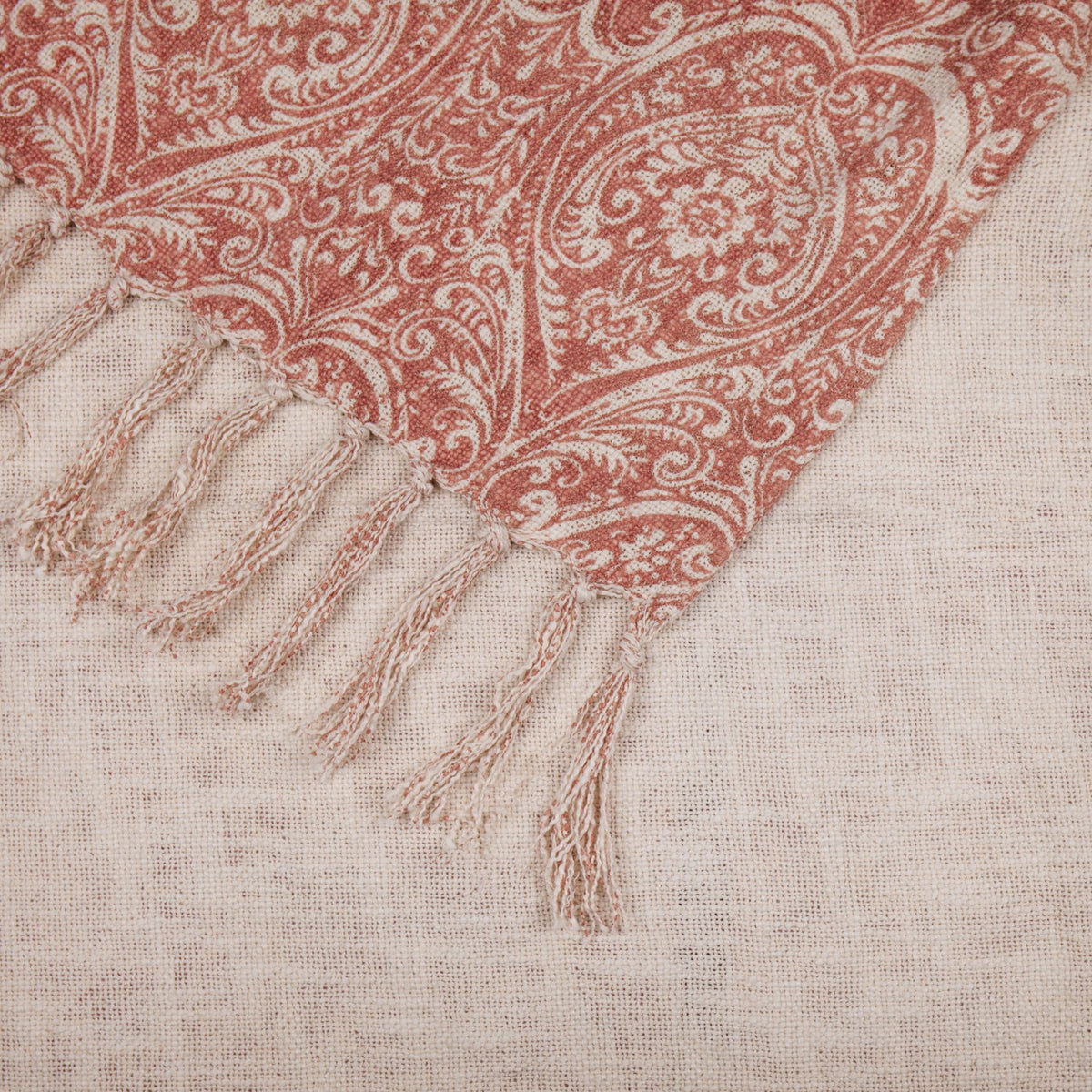 Corner detail of the Durelle Damask Cotton Throw in coral, showing the paisley and floral print edge with knotted tassel fringe against the plain cream reverse.
