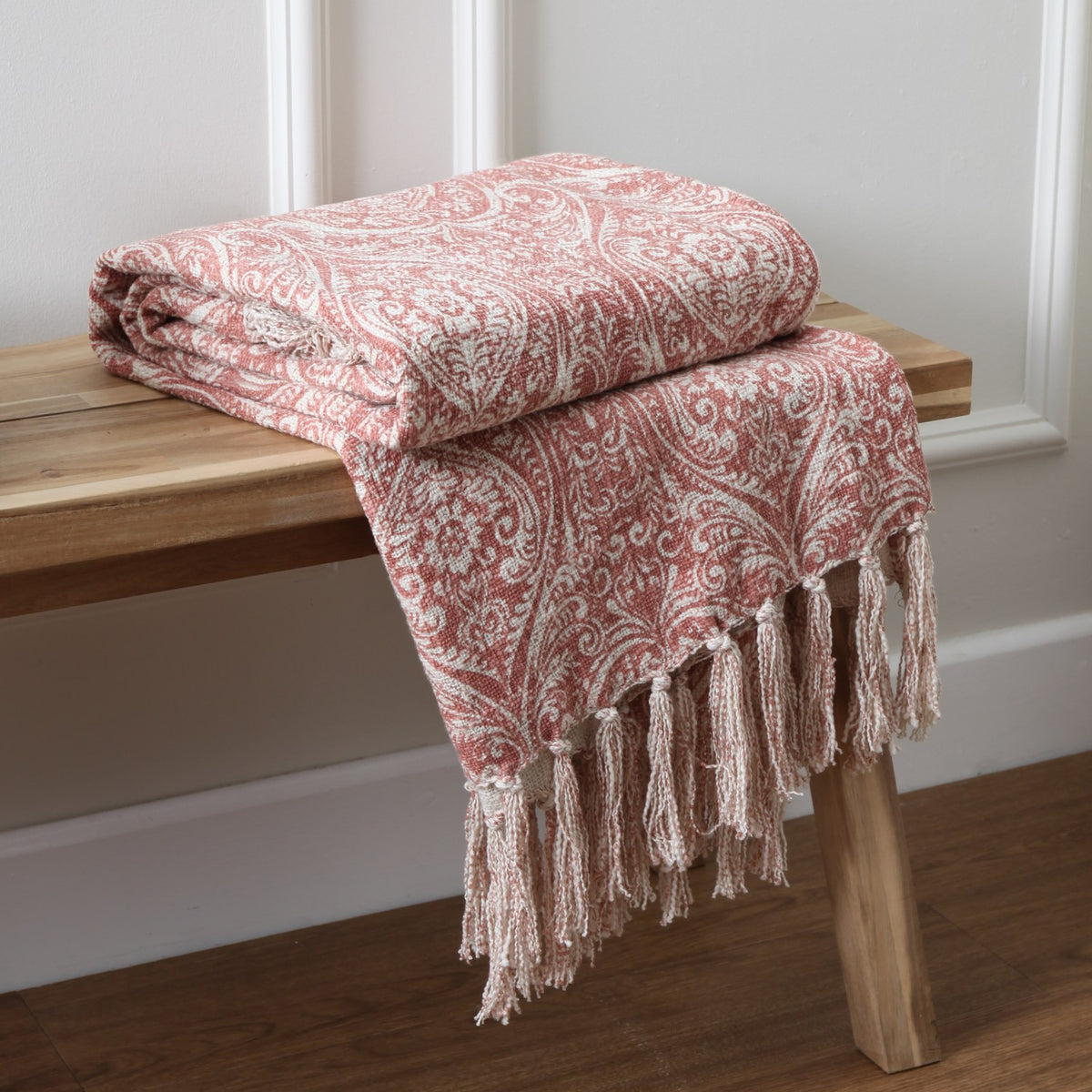Durelle Damask Cotton Throw in coral, folded on a wooden bench showing the paisley-floral pattern and knotted tassel trim against a white panelled wall.