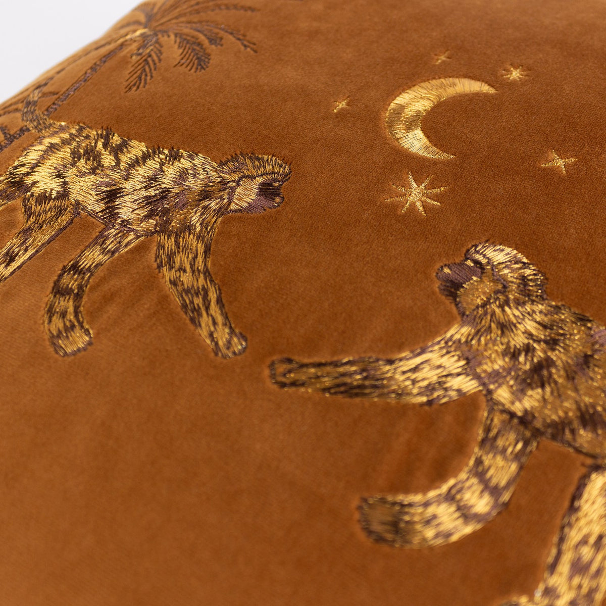 Close-up of the Dusk Animal Velvet Cushion in rust, showing embroidered monkeys, palm trees, and a crescent moon with stars stitched in gold and brown thread on velvet.