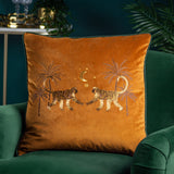 Dusk Animal Velvet Cushion in rust, embroidered with facing monkeys, palm trees and a gold crescent moon design, shown on a green velvet armchair.