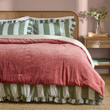 Edie Linen Cotton Bedspread in Rose, a quilted rose pink velvet throw with tied stitch detailing, styled on a bed with green striped pillowcases and a cane headboard in a bedroom setting.