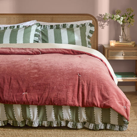 Edie Linen Cotton Bedspread in Rose, a quilted rose pink velvet throw with tied stitch detailing, styled on a bed with green striped pillowcases and a cane headboard in a bedroom setting.
