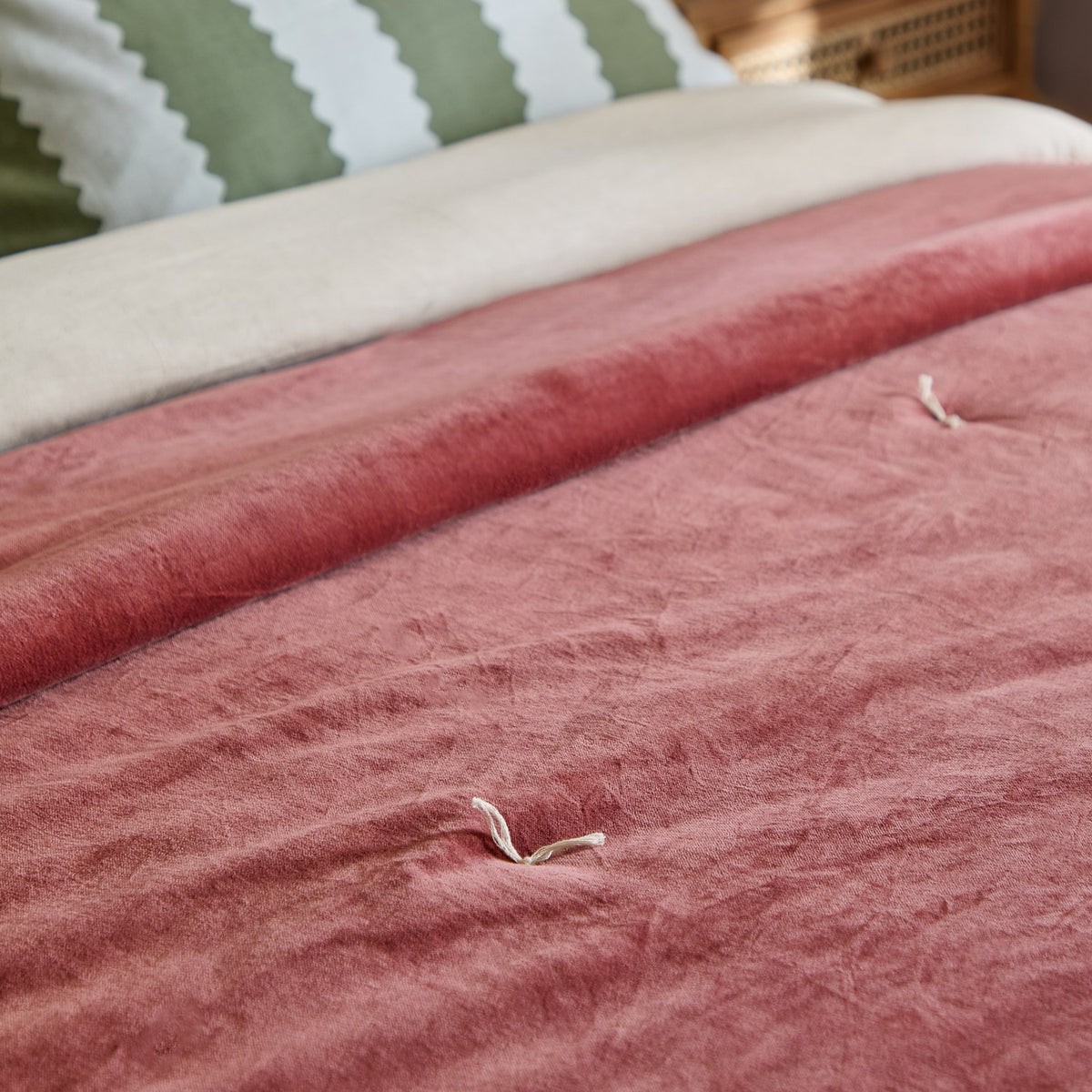 Edie Linen Cotton Bedspread in Rose, rolled on a bed to show its quilted cotton velvet face with tied stitch details, styled against green striped pillows.
