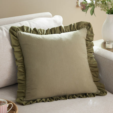 Effie Plain Cotton Cushion in sage green with a gathered ruffle trim, shown on a beige sofa in a living room setting.