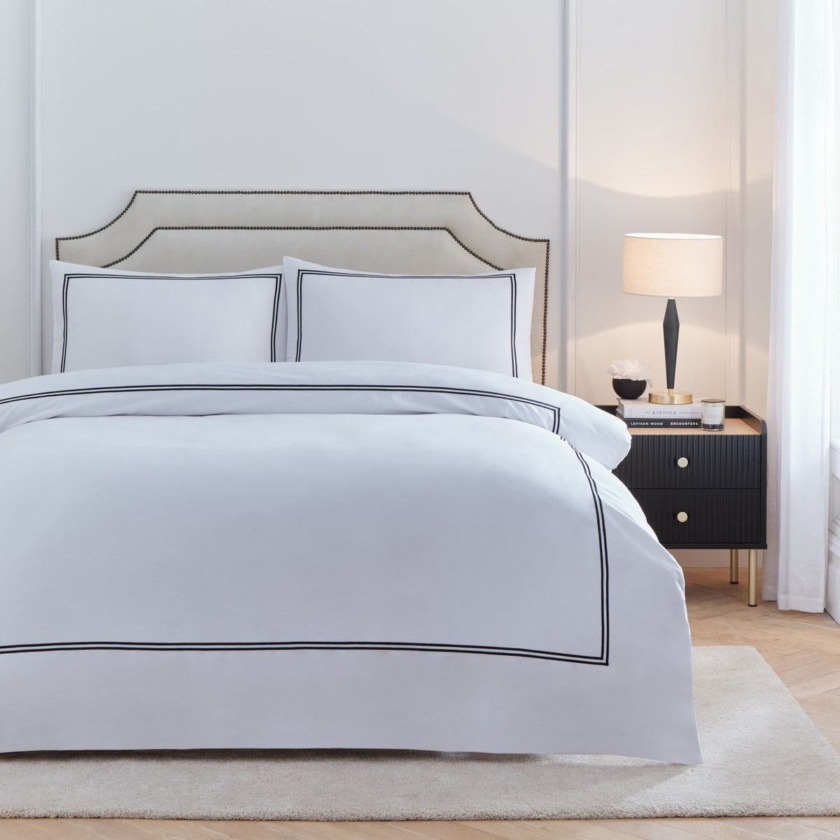 Elara Embroidered Cotton Duvet Cover Set in white with black twin-line border, shown with matching pillowcases on a bed in a bedroom setting with upholstered headboard and dark bedside table.