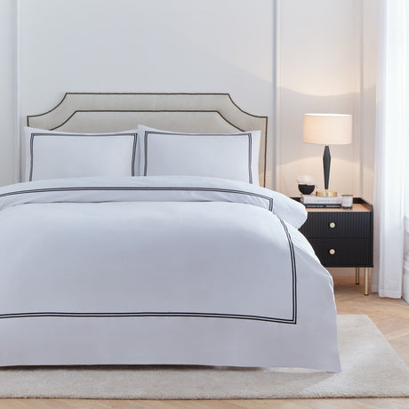 Elara Embroidered Cotton Duvet Cover Set in white with black twin-line border, shown with matching pillowcases on a bed in a bedroom setting with upholstered headboard and dark bedside table.
