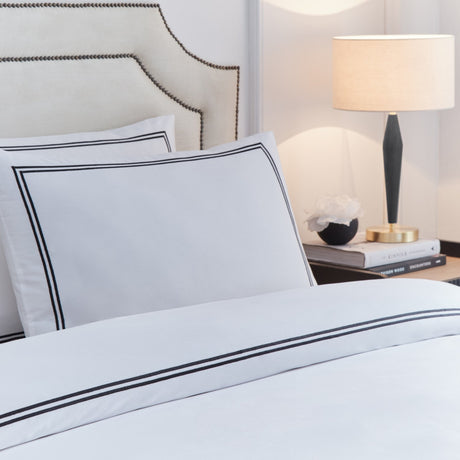 Elara Embroidered Cotton Duvet Cover Set in white with black twin-line border, shown on a bed with matching pillowcase in a bedroom setting beside a table lamp.