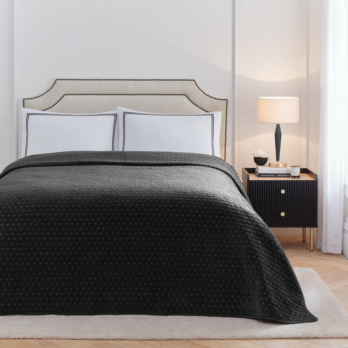 Bedroom setting with a black quilted Elara bedspread on a bed with white embroidered-border pillowcases, upholstered headboard, and black bedside table with lamp.