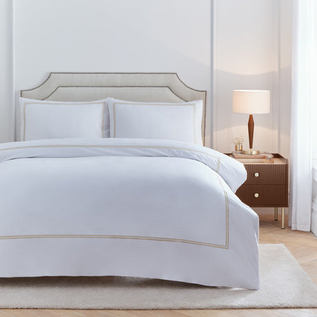 Elara Embroidered Cotton Plain Duvet Cover Set in white with champagne twin-line stitched border, shown with matching pillowcases on a bed in a bedroom setting.