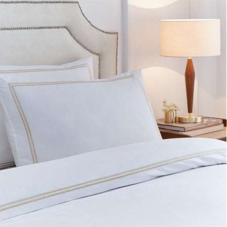Elara white cotton duvet cover set with champagne double-line embroidered border, shown on a bedroom bed with matching pillow shams beside a wooden table lamp.