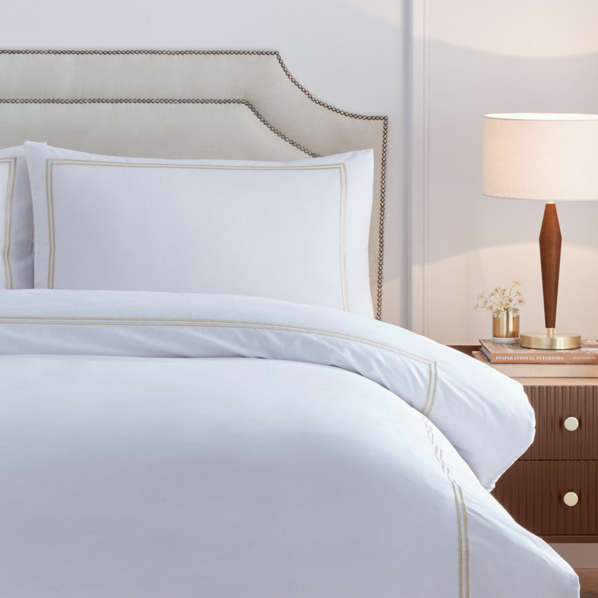 Elara Embroidered Cotton Duvet Cover Set in white with champagne double-line stitched border, shown with matching pillowcase on a bed in a bedroom setting.