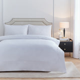 Elara Embroidered Cotton Plain Duvet Cover Set in white on a bed with matching pillowcases, shown in a bedroom with a beige upholstered headboard and black bedside table.