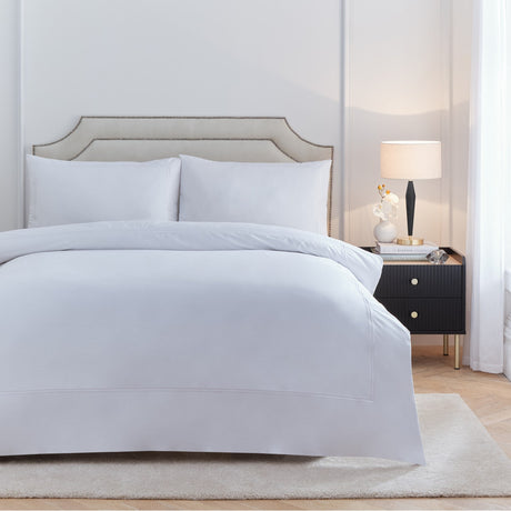 Elara Embroidered Cotton Plain Duvet Cover Set in white on a bed with matching pillowcases, shown in a bedroom with a beige upholstered headboard and black bedside table.