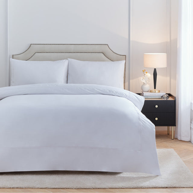 Elara Embroidered Cotton Plain Duvet Cover Set in white on a bed with matching pillowcases, shown in a bedroom with a beige upholstered headboard and black bedside table.