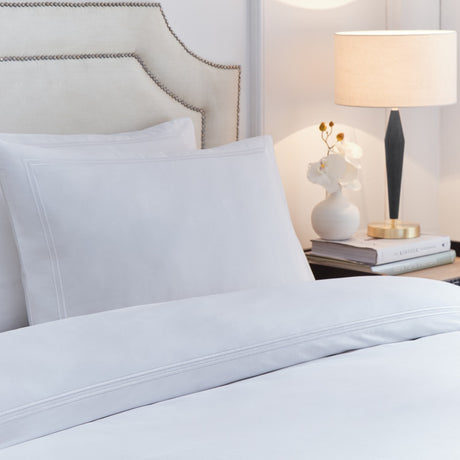 Elara white cotton duvet cover set with embroidered double-line border on cover and pillowcases, styled on a bed beside an upholstered headboard and bedside lamp.